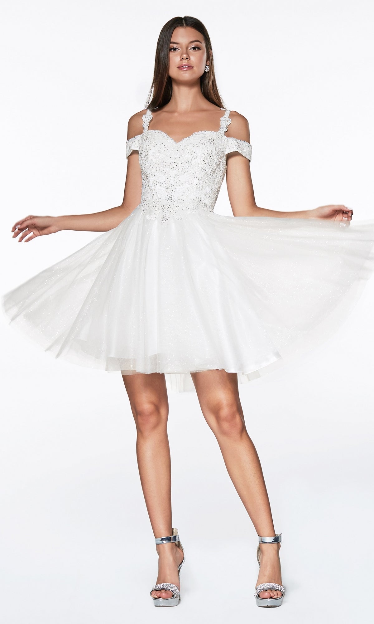 A woman models the Cold-Shoulder Short Glitter Homecoming Dress CD0132, featuring off-the-shoulder lace and sequin details, paired with silver high-heeled sandals. She holds out the flared sides to display its sparkling elegance.